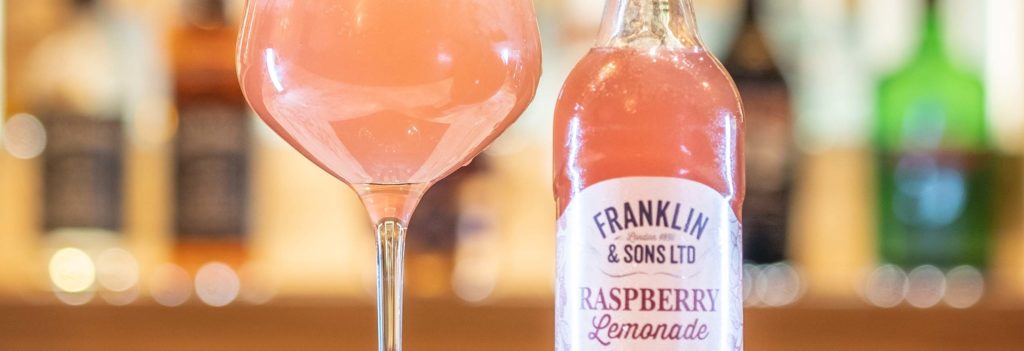 A wine glass filled with a pink cocktail next to franklin & sons raspberry lemonade