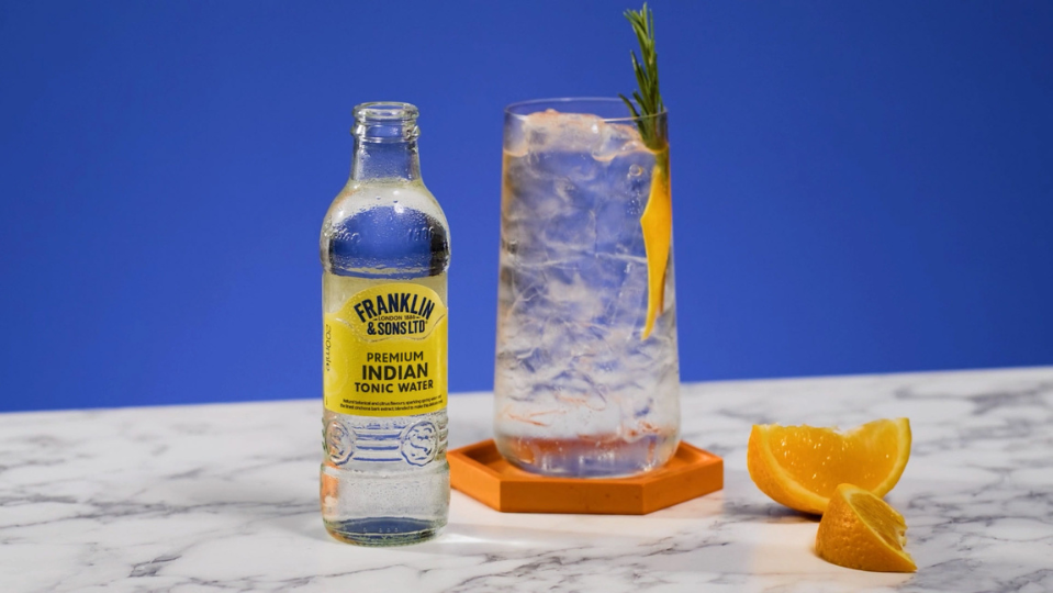 Franklin & Sons Premium Indian Tonic Water next to a clear cocktail with rosemary sticking out as the garnish.
